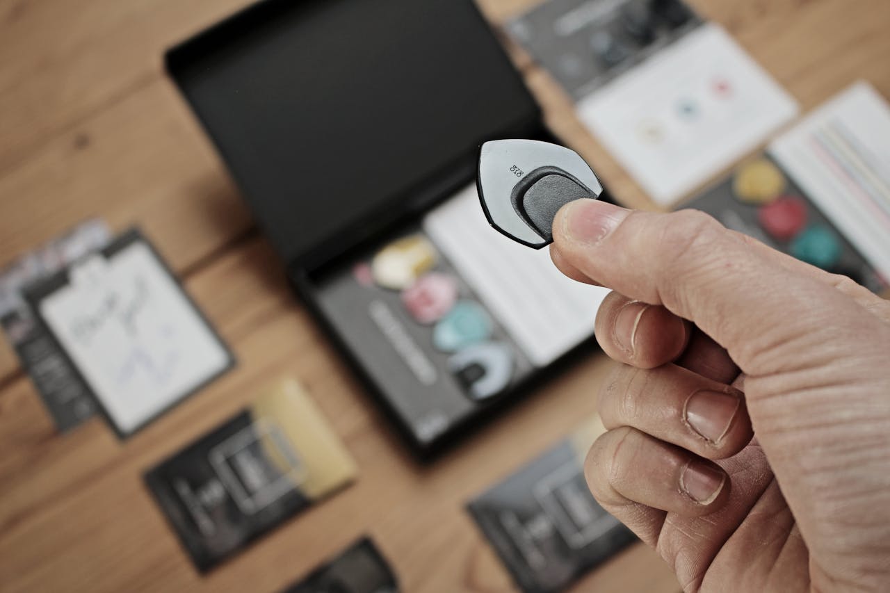 A close-up shot of a hand holding a guitar pick with a blurred background of a wooden table and packaging.