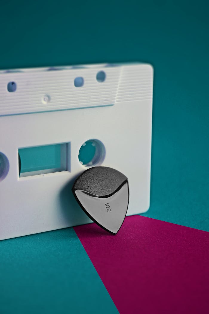 A close-up of a vintage cassette tape with a modern guitar pick on bold colored background.